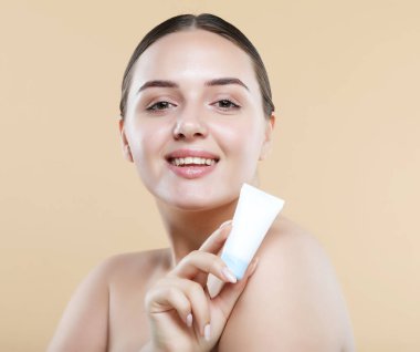 Beauty Youth Skin Care Concept - Beautiful Caucasian Woman Face Portrait holding and presenting cream tube product. Beautiful Spa model female with Perfect Fresh Clean Skin over beige background.