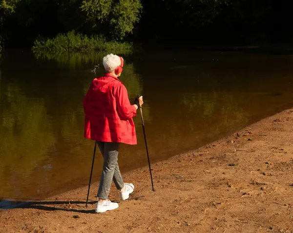 Elderly woman trains endurance while Nordic walking near lake . Old female listening music in headphones. Back view.