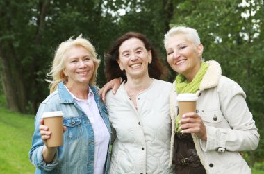 Lifestyle, emotion, old people and gold age concept. Charming elderly women friends are walking, talking and drinking coffee. Summer day in the park.