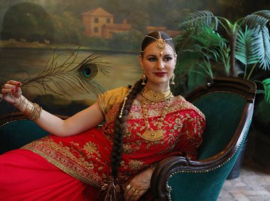Çekici genç bir kadın kanepede otururken tavus kuşu tüyü tutuyor. Geleneksel Hint düğün sarisi ve mücevherleri.