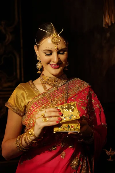 漂亮的年轻女子穿着传统的印度红莎丽和珠宝 手里拿着金棺材 美丽的女子对着深色的背景微笑 穆斯林 阿拉伯文化 — 图库照片