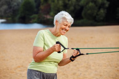 Gülümseyen yaşlı kadın spor giyer ve sabahları plajda egzersiz yaparken lastik bant kullanır. Sağlıklı yaşam tarzı, aktif emekli yaşam ve spor zamanı.