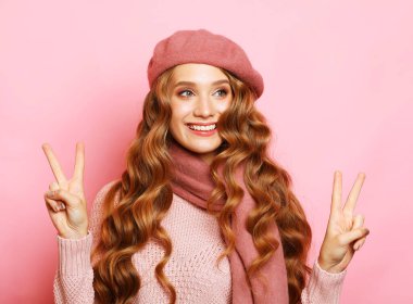 Yaşam tarzı ve insan konsepti: Sarı kıvırcık saçlı, pembe tişörtlü ve pembe arka planda bere takan genç, gülümseyen kadın. Kadın model v-işareti.
