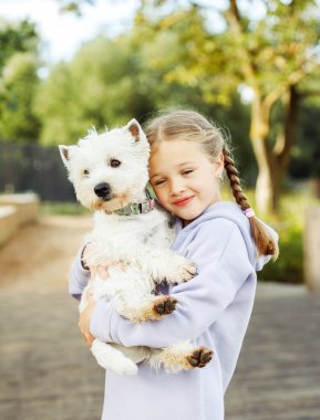 At kuyruklu küçük bir kız kollarında küçük beyaz bir köpek tutuyor ve mutlu bir şekilde gülümsüyor..