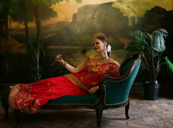 魅力的な若い女性がソファに座っている間クジャクの羽を持っています 伝統的なインドの結婚式のサリーとジュエリー — ストック写真