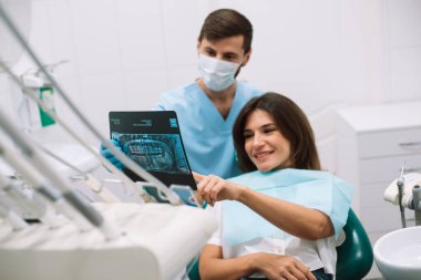 Diş hekimi çene röntgeni ve diş nakli gösteriyor. Orta yaşlı esmer kadın modern bir ofiste dişçi randevusunda.