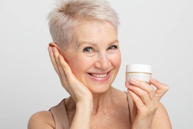 Gülümseyen 60 'lı yaşlarda olgun bir kadın yüzüne krem sürüyor açık gri arka planda kameraya bakıyor..