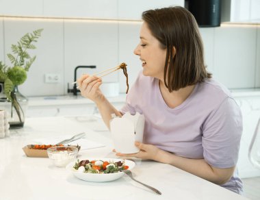 Orta yaşlı çekici kadın çubuklarla erişte yiyor, Kithcen 'de elinde karton kutu tutuyor.