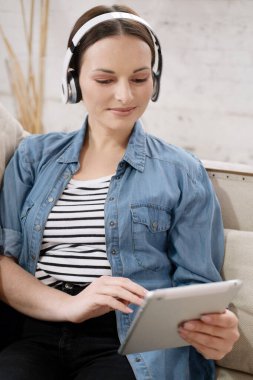 Rahat gülümseyen genç bir kadın evde koltukta otururken kulaklıkla film izliyor.