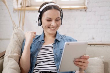 Rahat gülümseyen genç bir kadın evde koltukta otururken kulaklıkla film izliyor.