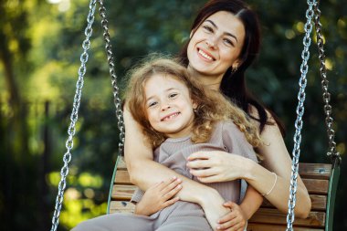 Genç anne ve 7 'nin kızı salıncakta sallanıyor. Beyaz kadın ve küçük kız oyun alanında eğleniyor..