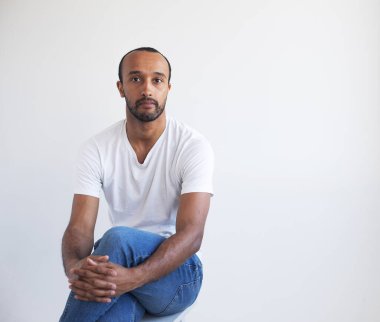 Beyaz tişörtlü, gri arka planda izole bir sandalyede oturan genç afro adamın fotoğrafı.