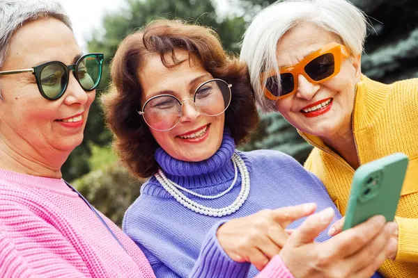 Three cheerful elderly friends in bright sweaters take a selfie on a smartphone and laugh, having a great time. Happy pensioners. Summer in the city.