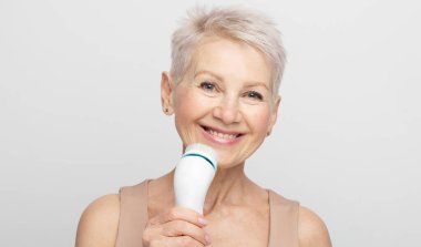 Gülümseyen, kısa saçlı yaşlı kadın mükemmel cilt için yüz masajı aleti kullanıyor.