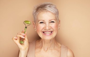Gülümseyen, kısa saçlı yaşlı kadın bej arka planda mükemmel bir cilt için yüz masajı yapıyor.