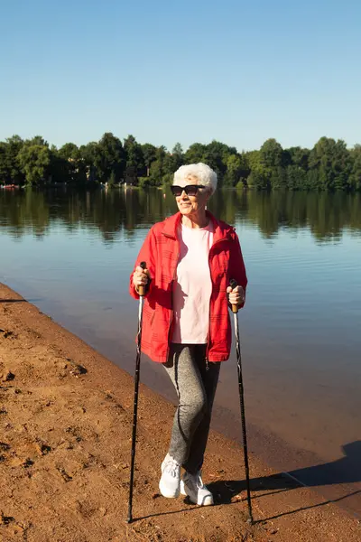 一位白发苍苍的老妇人 戴着太阳镜 身穿红色夹克 手持跟踪棒行走在湖边的沙滩上 — 图库照片