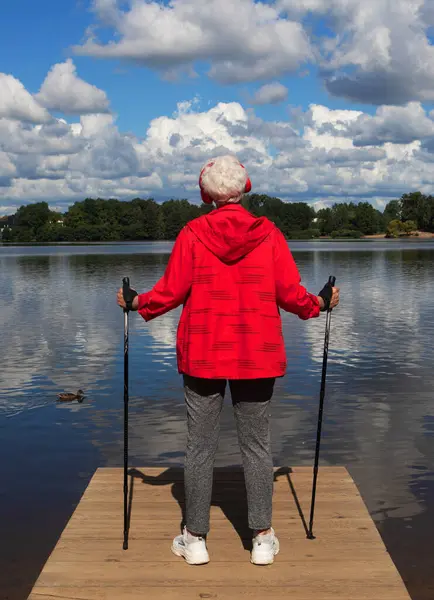 An elderly woman with short hair, dressed in a red jacket, stands with Nordic walking poles on a bridge by the lake nd listening music with headphones.
