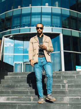 Genç ve yakışıklı Afro adam güneş gözlüğü takıyor. Modern iş merkezinin yakınına mesaj göndermek için akıllı telefon kullanıyor..