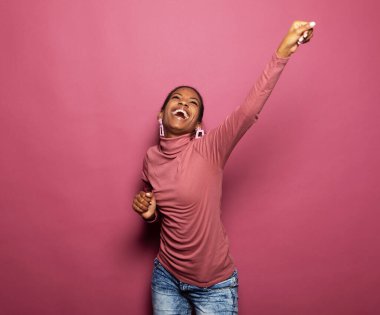 Mutlu gülümseyen genç afro kadın pembe arka planda tek başına dans ediyor.