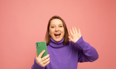 Yetişkin bir kadın zaferi kutluyor. Bir yumruğunu havada tutuyor. Pembe arka planda izole bir süveter giyiyor.