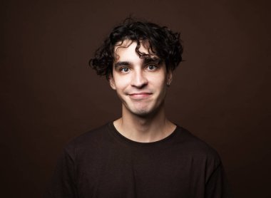 Yakışıklı genç adam kahverengi tişört giyer ve kahverengi arka planda kameraya bakar. Portreyi kapat.