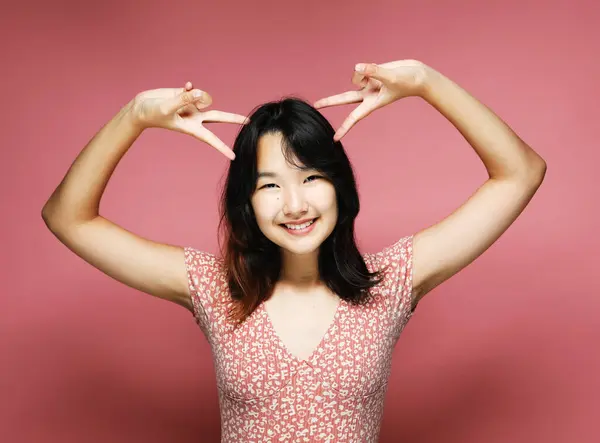 Joven Sonriente Asiático Mujer Usa Rosa Vestido Mostrando Victoria Signo Imagen De Stock