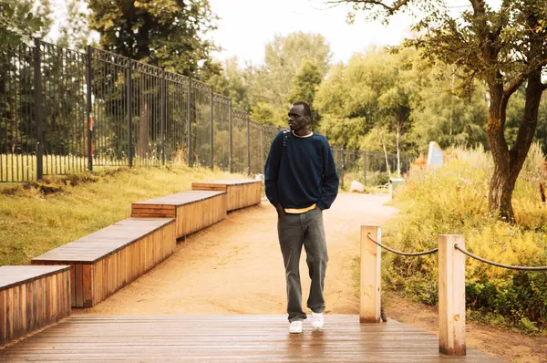 青いスウェットシャツを着た若いハンサムなアフリカ人男性の肖像画 バックパックを持ち 夏の日に公園を歩く ストック写真