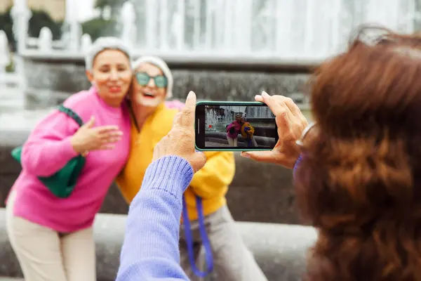 Three cheerful elderly friends in bright sweaters take a photo on a smartphone and laugh, having a great time.