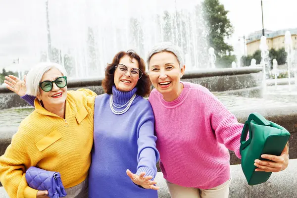 三个穿着鲜艳毛衣和太阳镜的快乐的退休女性朋友走在一起 生活方式和人的概念 免版税图库照片
