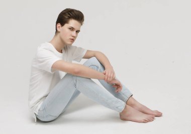 Çıplak ayaklı, genç, kot pantolonlu ve beyaz tişörtlü bir erkek model yerde oturuyor..