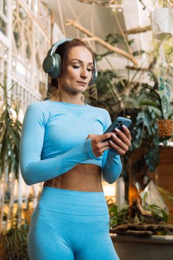 Akıllı telefon tutan ve kulaklık takan bir kadın müzik dinliyor, pilates yaparken dinleniyor, tropikal bitkilerle dolu bir odada yoga eğitimi alıyor..