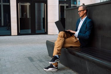 Young stylish man working on laptop computer while sitting on bench outdoors near modern office. Summer day.