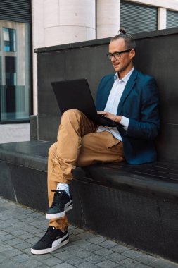 Young stylish man working on laptop computer while sitting on bench outdoors near modern office. Summer day.