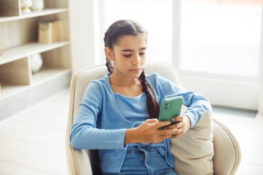 Küçük tatlı kız sandalyede oturuyor, evde eğleniyor, akıllı telefon kullanıyor, cep telefonuyla video oyunu oynuyor..