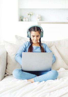Örgü saçlı, kulaklık takan kız öğrenci kanepede oturur ve dizüstü bilgisayarla çalışır.