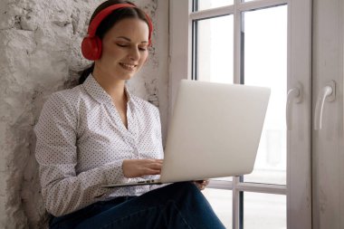 Evde dizüstü bilgisayar ve kırmızı kulaklıklarla oturan genç bir kadın.