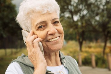 Mutlu yaşlı kadın yaz parkında akıllı telefonuyla konuşuyor ve gülümsüyor..