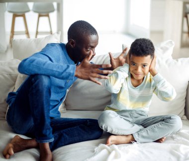 Kızgın baba evde dikilmiş ona bağırırken siyah küçük çocuk kulaklarını tıkıyor. Aile içi şiddet ve mutsuz çocukluk..