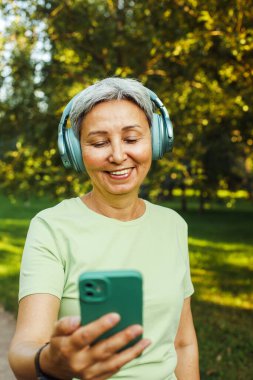 Mutlu ve gülümseyen 60 yaşındaki Asyalı kadın yeşil parkta yürürken torunuyla video görüşmesi yapıyor. Hobi ve eğlence konsepti.