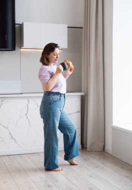 Kapalı alanda, pencerenin yanında, mutfakta kruvasanla kahve içen sevimli, kıvrımlı esmer kadın resmi..