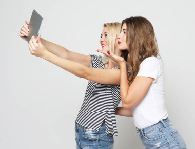 Gülümseyen bayan arkadaşlar beyaz arka planda dijital tabletle selfie çekiyorlar..