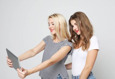 Gülümseyen bayan arkadaşlar beyaz arka planda dijital tabletle selfie çekiyorlar..