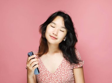 Genç, güzel, Asyalı bir kadın gülümseyip yıllıklarla müzik dinliyor. Elinde pembe bir arka planda cep telefonu tutuyor.,