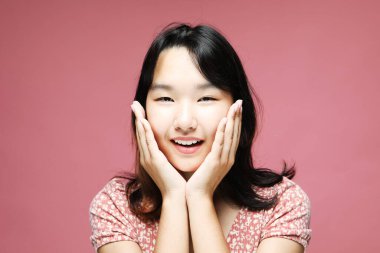 Elleri yanaklarında genç bir Asyalı kadın, kameraya bakıyor, sağlam bir arkaplan, portre fotoğrafı, pembe tonlar, orta çekim, yüksek çözünürlüklü detaylar..