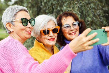 Üç orta yaşlı kadın parkta selfie çekiyor, renkli ve güneş gözlüğü takıyorlar. Akıllı telefonla fotoğraf çektirirken gülümsüyor. Sevimli yaşlı arkadaşlar dışarıda birlikte eğleniyorlar..