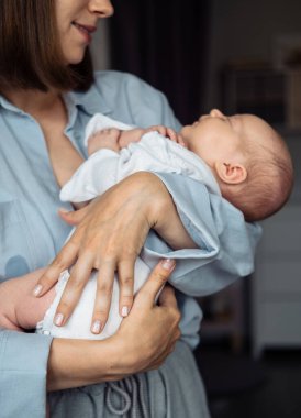 Yeni doğmuş bebeği kucağında taşıyan genç bir annenin yakın plan portresi. Evde mavi gömlek giyiyor. Yeni doğmuş çocuğa sarılırken kameraya poz veren genç kadın..