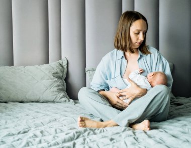 Açık mavi pijamalı bir anne yeni doğmuş bebeğini iki eliyle tutarak gri yoga pantolonu giyer. Arka plan gri-mavi, geniş bir açıklık ve yumuşak ışıklandırmayla. Sıcak renkler ve doğal hareketler çocuklara olan sevgiyi gösteriyor.