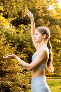 Parkta yoga yapan genç bir kadın, dışarıda yaz günü spor yapan güzel bir kızın portresi. Genç bir bayan, açık mavi bir spor giyerken ve doğal bir arka planda otururken mükemmel bir vücut ve güzel bir yüzle esniyor..