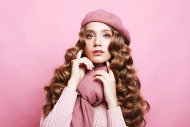 Pembe arka planda pembe kazak, eşarp ve bere giyen kıvırcık saçlı genç bir kadın..