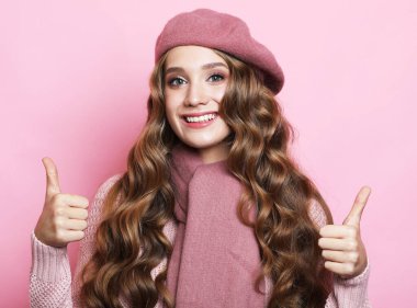 Pembe arka planda pembe kazak, eşarp ve bere giyen kıvırcık saçlı genç bir kadın..
